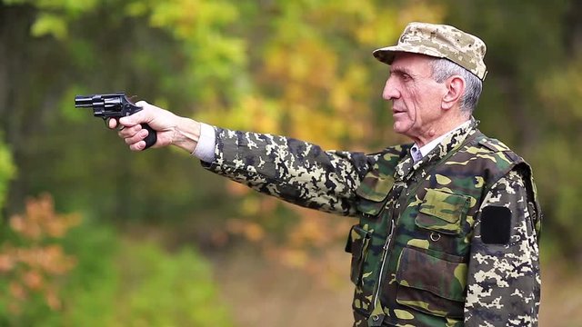 Elderly person in military uniform shoots a revolver. Retired officer at shooting range. Senior man in military uniform shoots a pistol in forest. Man with black gun. Video with sound
