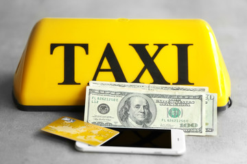 Yellow taxi roof sign with phone, credit card and American dollars on gray background, closeup