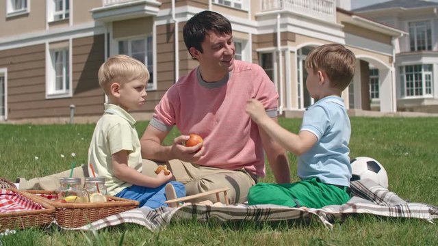 Father having picnic on green lawn with two little sons and smiling while one of them talking about something to him
