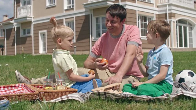 Man sitting on green lawn with two little sons and smiling while them explaining and showing him something funny
