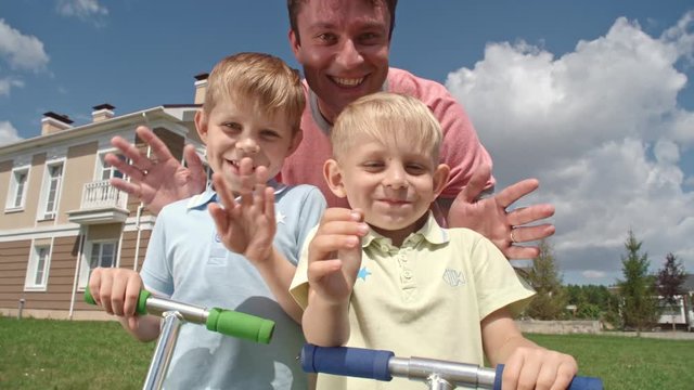 Father and two hilarious boys holding handlebars of scooters, smiling, making funny faces and waving at camera