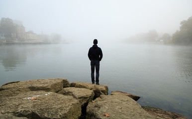 Fototapeta na wymiar Man standing at the shore of the Saone and looking over the river during a foggy, autumn morning.