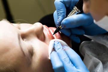 Cosmetologist applying permanent make-up. Young beautiful woman having cosmetic tattoo on her lips. Healthy Spa