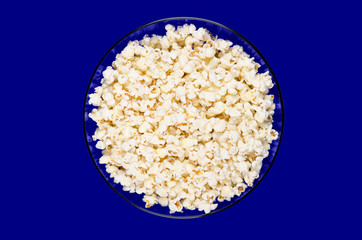 Popped popcorn in a blue glass bowl on blue background. Butterfly shaped popcorn puffed up from the kernels, after it has been heated. Edible and vegan food. Isolated macro photo close up from above.