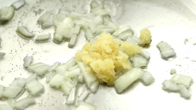 Crushed garlic added to fry pan with hot oil and onion being cooked and stirred, closeup slow motion