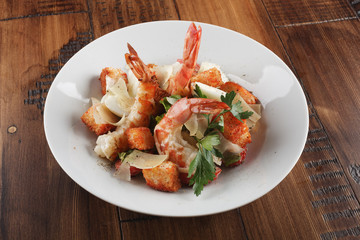 Caesar salad with king prawns in a white plate. Salad with shrimp on wooden brown background.
