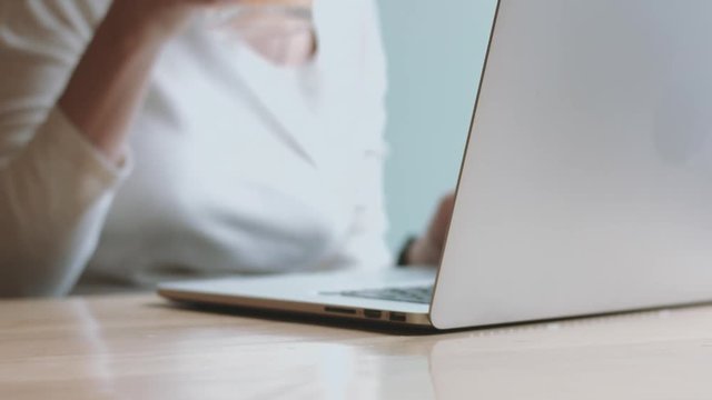 Cropped faceless woman sitting at table using laptop and drinking juice from faceted glass near her.