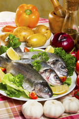 Three pieces of raw and fresh fish, sea bream, sea bass and trout on a white plate beside fresh vegetables and lemon.