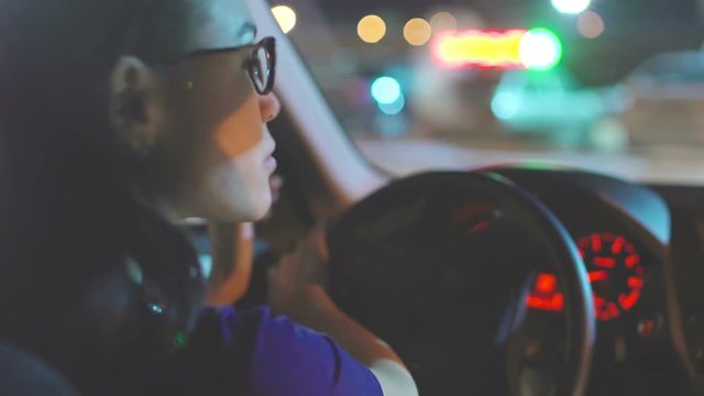 Close up of young and beautiful Asian woman in glasses driving car through shadowy night city