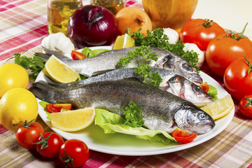 Three pieces of raw and fresh fish, sea bream, sea bass and trout on a white plate beside fresh vegetables and lemon.