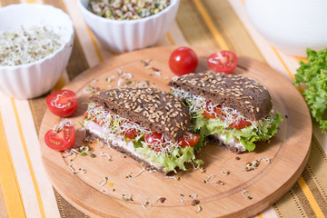 A healthy meal of whole wheat dark (brown) rye bread, curd cream cheese, radish, alfalfa sprouts, ham, cherry tomatoes and green salad on a wooden cutting board. Delicious breakfast full of vitamins.