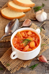 Cabbage soup with meat in a bowl, spoon, garlic, dill, pieces of bread on old wooden background. Soup cooked with meat, potatoes, cabbage, carrots, garlic, onion and tomato. Rustic style