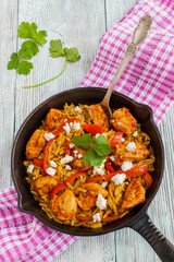 One-pot chicken fillet and orzo pasta with red bell peppers and feta cheese, cooked with garlic, paprika and olive oil. Cast-iron skillet and fresh parsley on wooden table, top view.