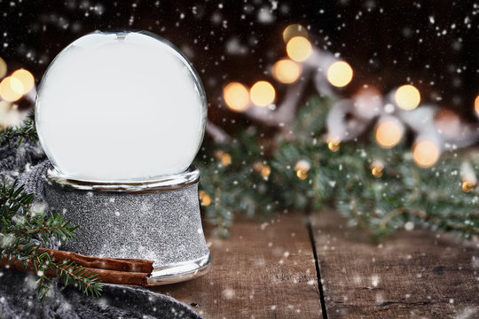 Rustic image of an empty Christmas snow globe surrounded by pine branches, cinnamon sticks and a warm gray scarf with copy space and gently falling snow. Selective focus on snowglobe.