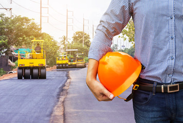 engineer holding safety helmet at road construction site with roller compactor working asphalt road