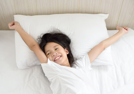 Portrait of happy young Asian girl lying in bed after waking up and stretching arms on the bed in the morning after good night sleep. Cheerful kid model with dreamy expression relaxing in the morning.
