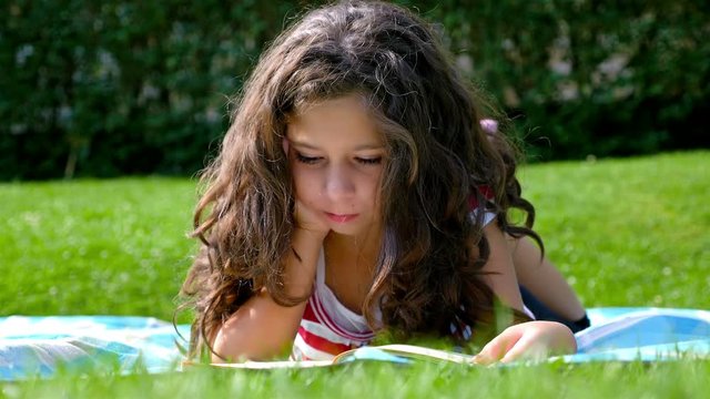 Cute little girl with curly hair is reading a book in the garden