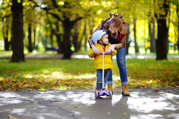 Young mother showing her toddler son how to ride a scooter in a autumn park