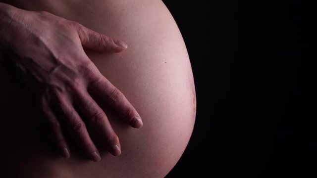 Pregnant woman touching caressing her naked big belly, closeup on black background. Pregnancy, motherhood and baby anticipation concept. 4K ProRes HQ codec