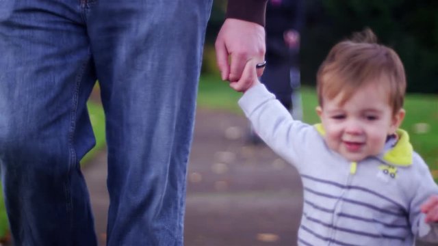 Adorable little boy holding his dad's hand while walking in a park on a fall day