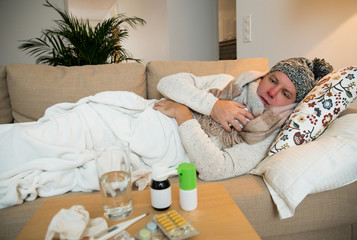 Sick man wearing scarf lying on couch at home under a blanket checking temperature. Living room with table full of medicine and pills. Man with running nose and cough. 