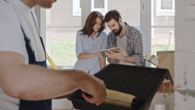 Closeup of contractor applying plaster to wall with paint roller in new house while couple sitting and discussing interior plan with digital tablet and paper model in the background