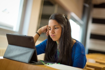 Bored young female student at university classroom.
She's using tablet and headphones for taking notes.