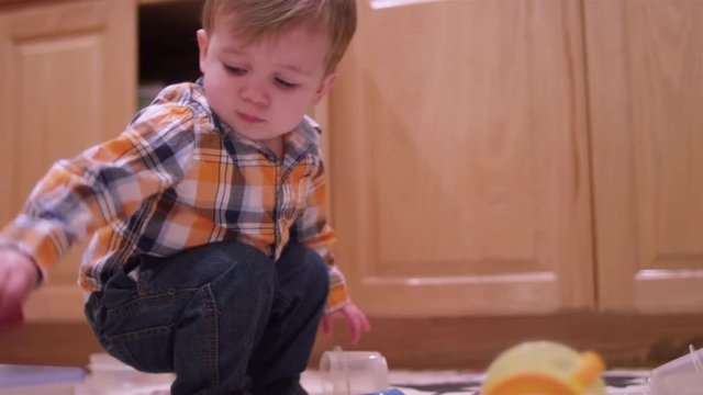 Little boy playing with plasticware in the kitchen and rubbing his eyes and looking tired