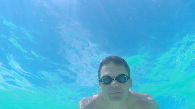 Slow motion 60 fps underwater low angle shot of muscular Caucasian man wearing goggles and swimming breast stroke under surface of sea water toward and over camera