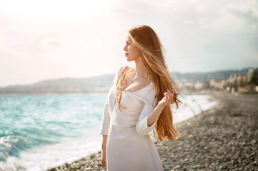 Fototapeta na wymiar Outdoor summer portrait of young pretty tourist woman with great hair looking to the ocean at europe beach, enjoy her freedom and fresh air, wearing stylish white dress.