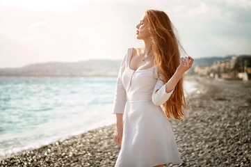 Outdoor summer portrait of young pretty woman with great hair looking to the ocean at europe beach, enjoy her freedom and fresh air, wearing stylish white dress.
