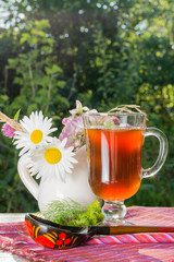 Бокал с квасом, деревянная ложка, зелень и кувшин с садовыми цветами на столе