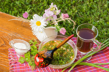 Тарелка с окрошкой, бокал с квасом, букет цветов в кувшине, соус и зелень на столе