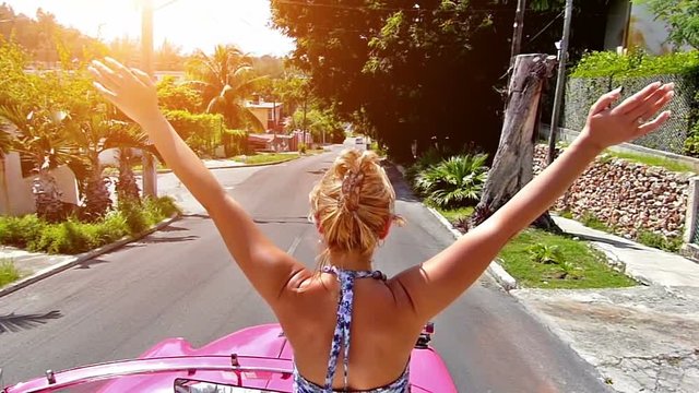 Rear view of happy blonde woman with arms outstretched, driving in pink cabriolet through Havana city, Cuba.Slow motion, high speed camera, lens flare