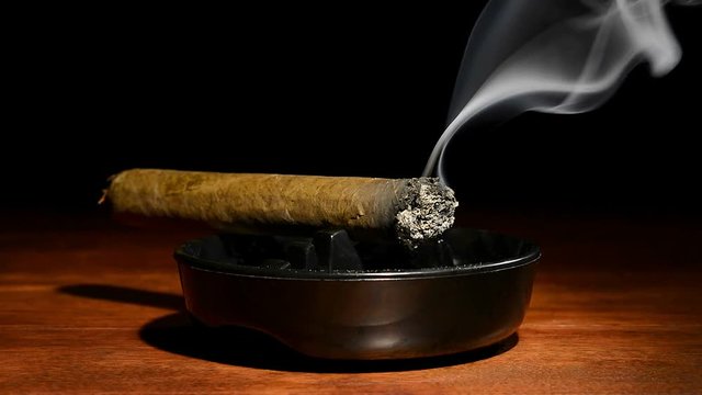 A burning cigar in a classic black ashtray streaming smoking in a dark, moody setting.