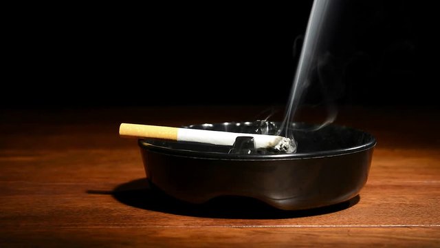A burning cigarette in a classic black ashtray streaming smoking in a dark, moody setting.