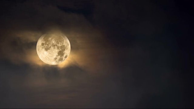 Fog moves across a full moon framed against a dark moody, amber sky with cloud cover..