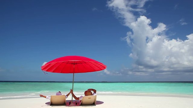 Rear view of happy couple with arms outstretched lying on back in deckchairs under the red parasol.White sand beach and turquoise water in Maldives beach