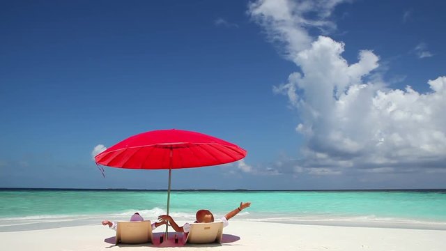 Rear view of happy couple with arms outstretched lying on back in deckchairs under the red parasol.White sand beach and turquoise water in Maldives beach