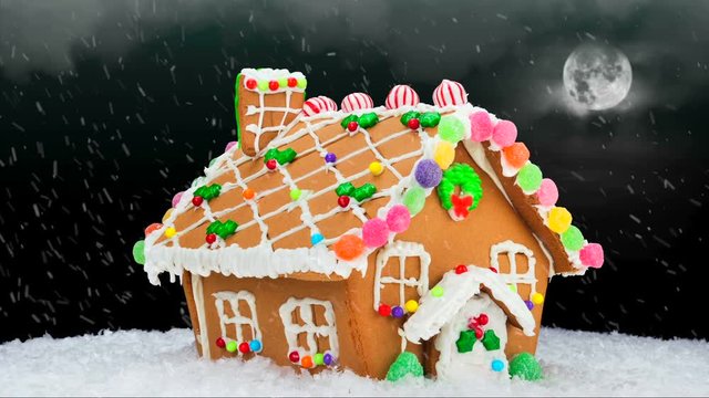 A gingerbread house during a cold, snowy Christmas evening shows a festive setting with a cloudy, black sky, bright moon and delicious cookie decoration..