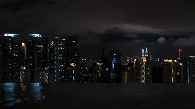 Timelapse shot of clouds sailing over night illuminated Kuala Lumpur, Malaysia. View from rooftop swimming pool. Rippling water in foreground