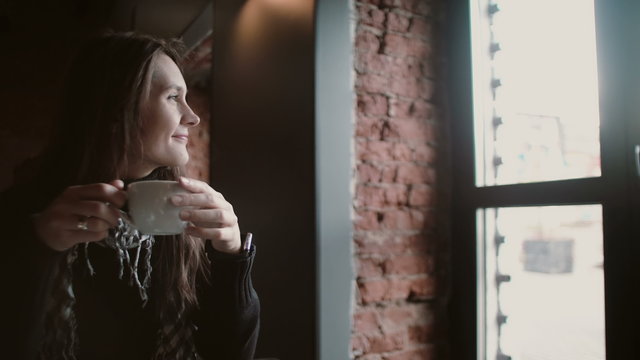 young girl drinking tea from a mug and looking out the window sitting in modern loft dining 4k