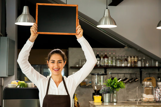 Beautiful Caucasian woman in barista apron holding empty blackboard sign inside coffee shop - ready to insert text