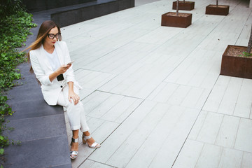 Young attractive businesswoman with long hair is using smart-phone sitting in a backyard near office building. Gray background with copy space area for your text o design.