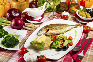 Fried gilt-head bream on a plate with a side dish of broccoli, cherry tomatoes, rice, olives and lemon served beside fresh vegetables and sauce.
