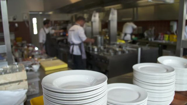 A pile of clean white plates with cooks busy in a restaurant kitchen in the background.
