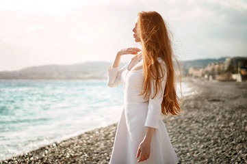 Outdoor summer portrait of young pretty woman with great hair looking to the ocean at europe beach, enjoy her freedom and fresh air, wearing stylish white dress.