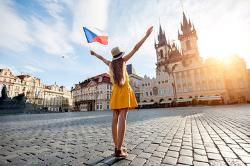Naklejka premium Young female tourist dressed in yellow holding czech flag on the old town square of Prague. Enjoying great vacation in Czech republic