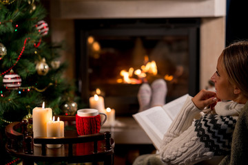 Woman is sitting with cup of hot drink and book near the fireplace