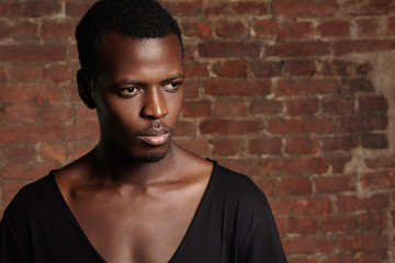 Fototapeta premium Indoor shot of young African male model with athletic body wearing black stylish T-shirt with scooped neck standing against red-brick wall with copy space for your text or promotional content